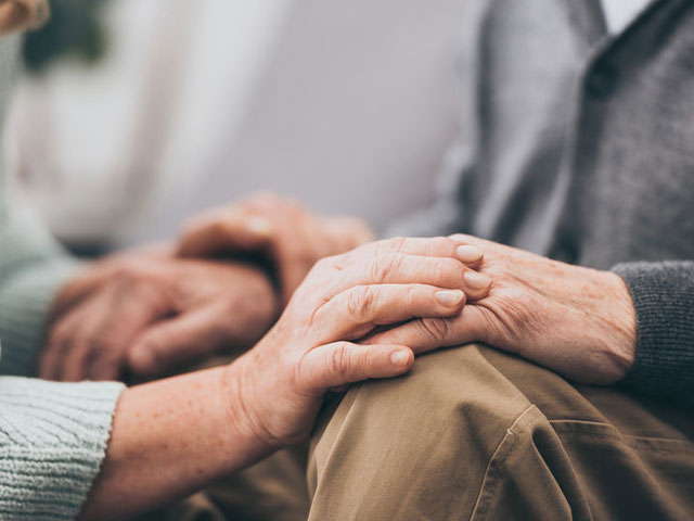 Two elderly people sit close together, holding hands for comfort and support.