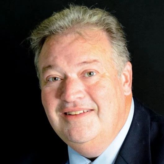 Middle-aged man with light hair wearing a dark suit and light blue shirt, smiling in front of a black background.